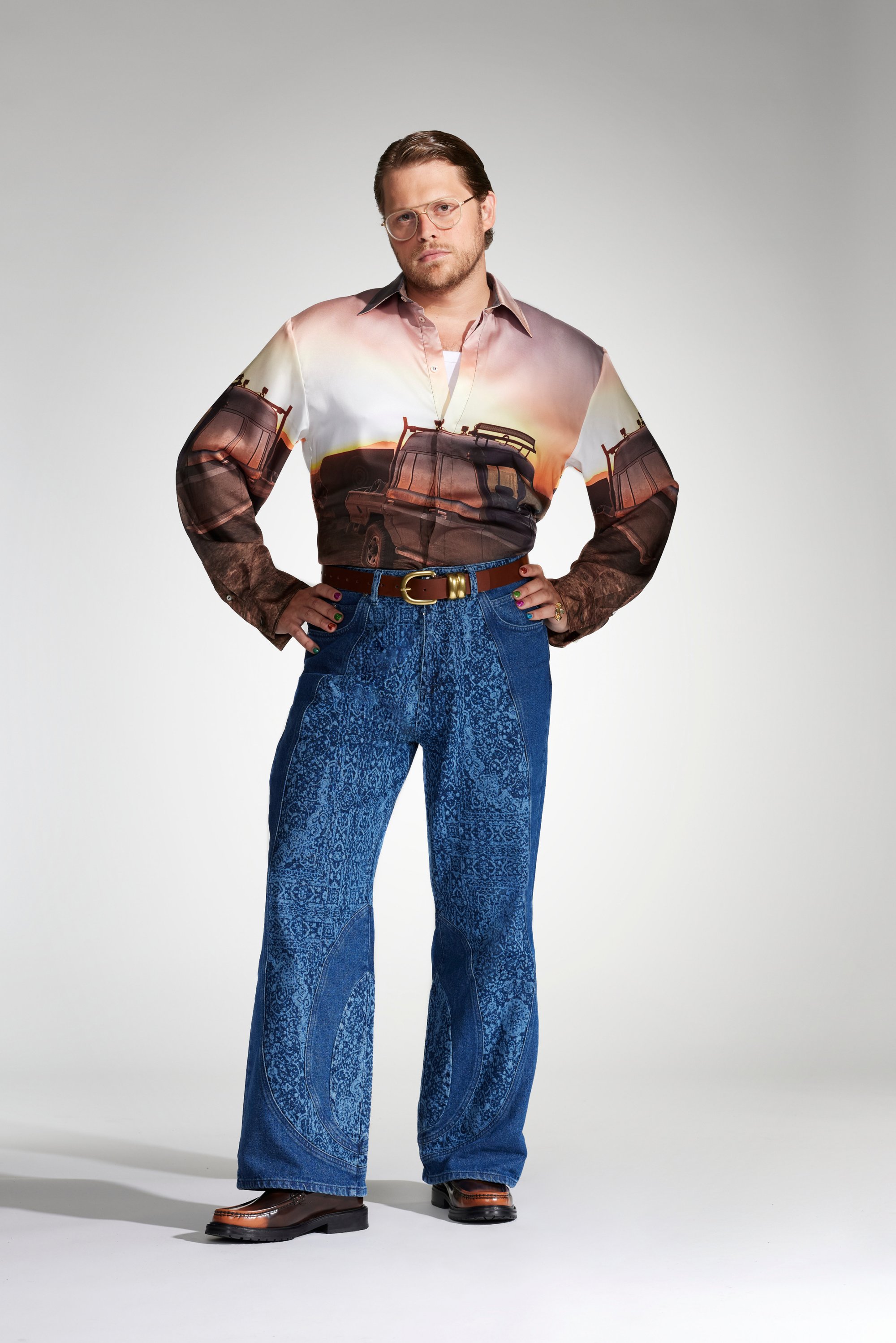 A man stands confidently, wearing a graphic shirt depicting vehicles and ornate blue jeans, complemented by stylish shoes and glasses.