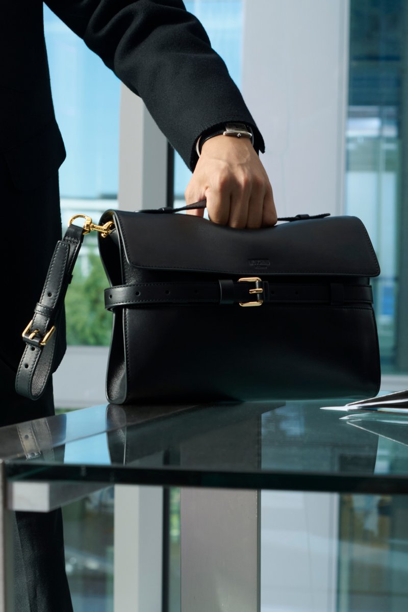 Borsa nera in pelle con patta, dettagli in metallo dorato, tracolla regolabile e chiusura simile a una cintura, appoggiata su una superficie in vetro.