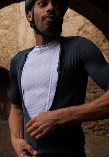 Un cycliste ajuste la fermeture éclair d'un maillot de cyclisme ajusté, mettant en valeur une couche inférieure texturée, sur un fond de pierre rustique.