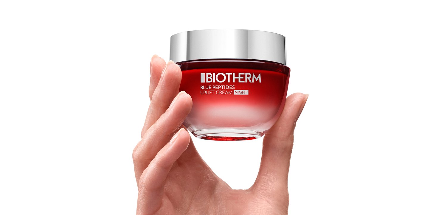 Red glass jar containing "Blue Peptides Uplift Cream Night" with a silver lid, held by a hand. Smooth texture, transparent base revealing cream inside.