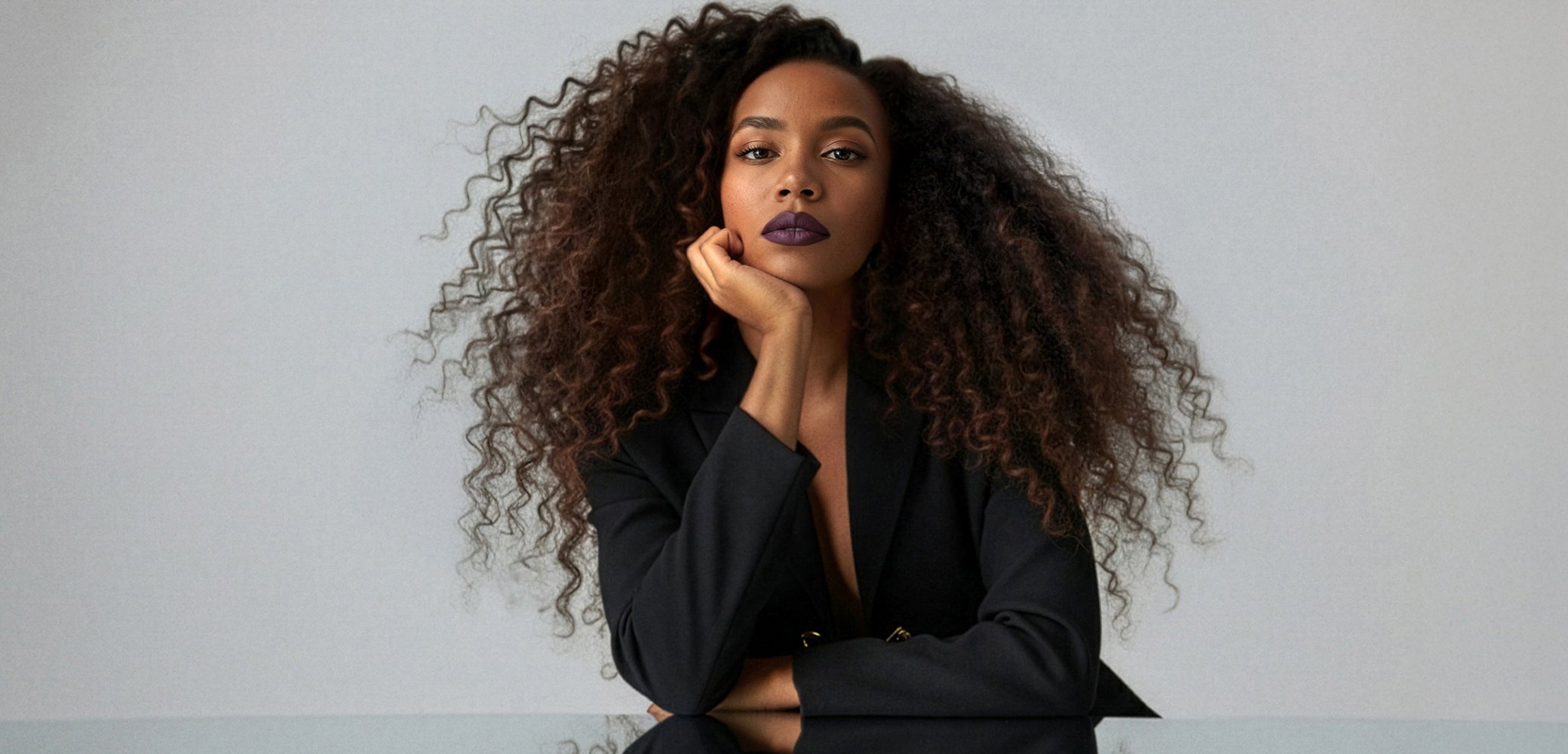Svart blazer med djup ringning, stort, voluminöst lockigt hår, mörkt läppstift. Slät yta i förgrunden, neutral bakgrund.
