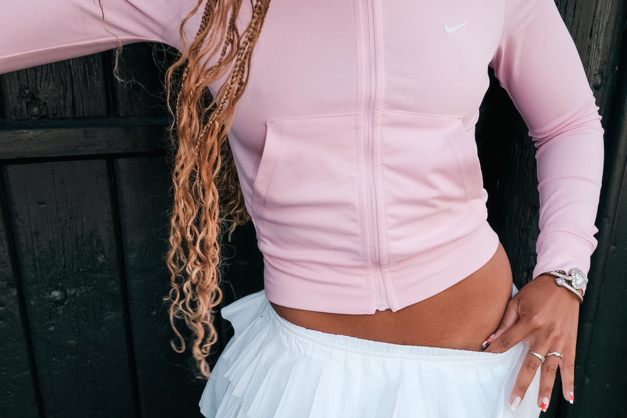 Persoon in een roze zip-up jas en een witte geplooide rok, met de hand die de tailleband van de rok vastpakt, lange krullende haren en sieraden om pols en vingers.