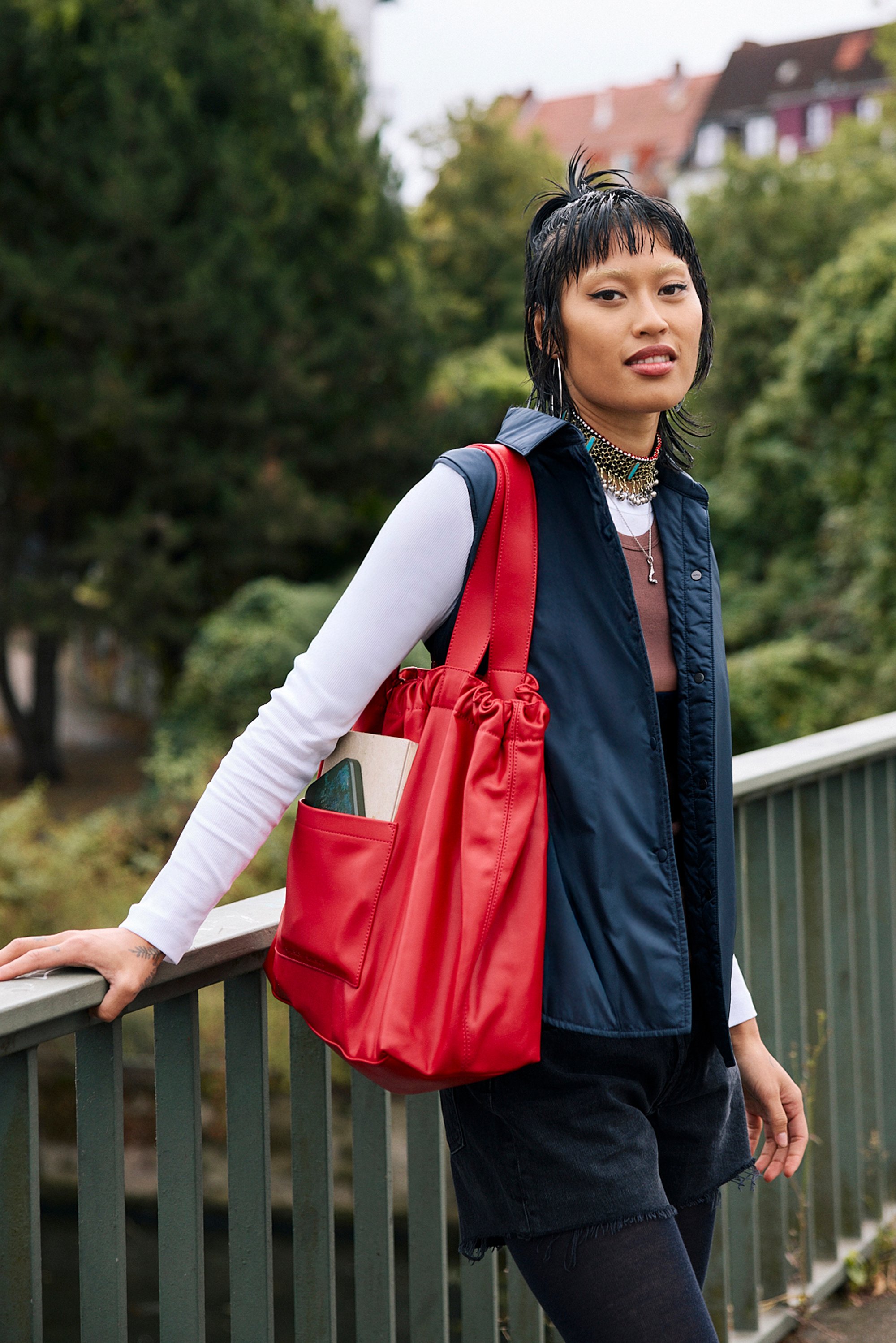 Red drawstring tote bag with a smooth texture. Worn with a navy sleeveless vest, white long-sleeve top, and dark shorts, set against lush greenery.