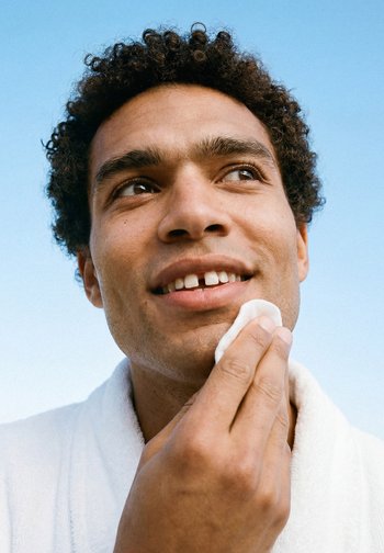 Homme aux cheveux bouclés souriant doucement en tenant un coton sur sa joue, avec un ciel bleu clair en arrière-plan.