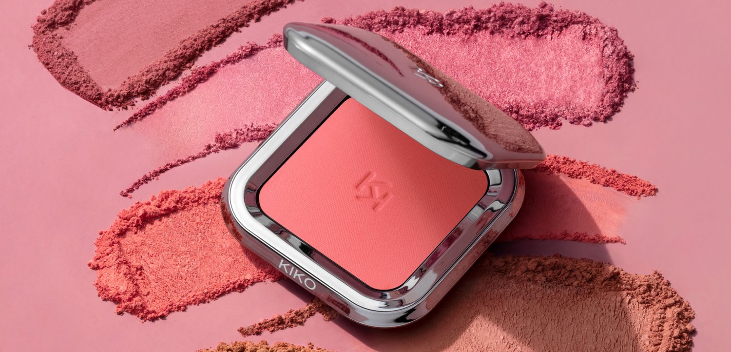 Kompakt pink blush med metallisk sølv låg delvist åbent, placeret på en overflade med forskellige pink og koral pulverprøver.