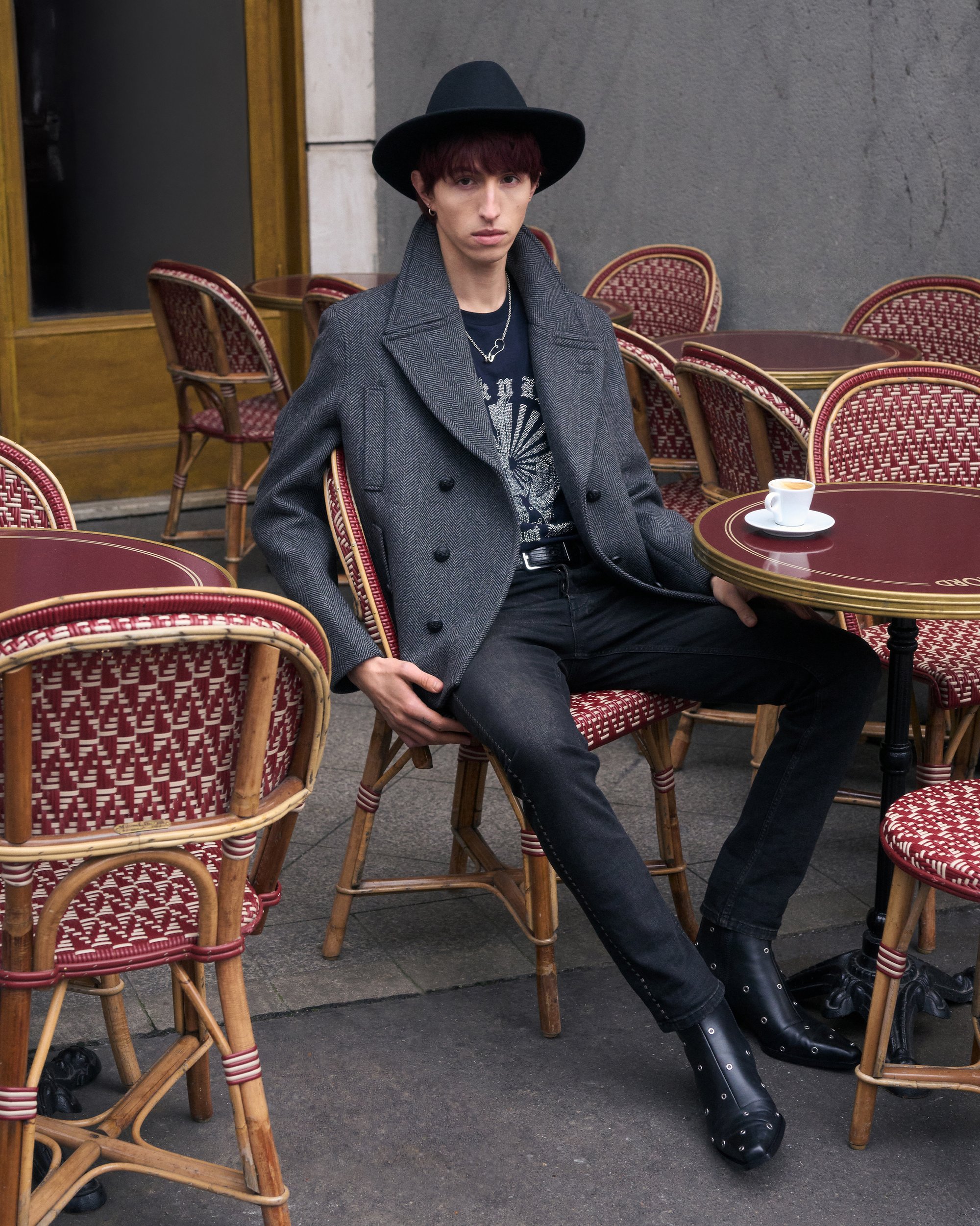 Una persona joven con el pelo corto está sentada en una mesa de café, vistiendo un abrigo gris y un sombrero negro, con una taza de espresso frente a ellos.