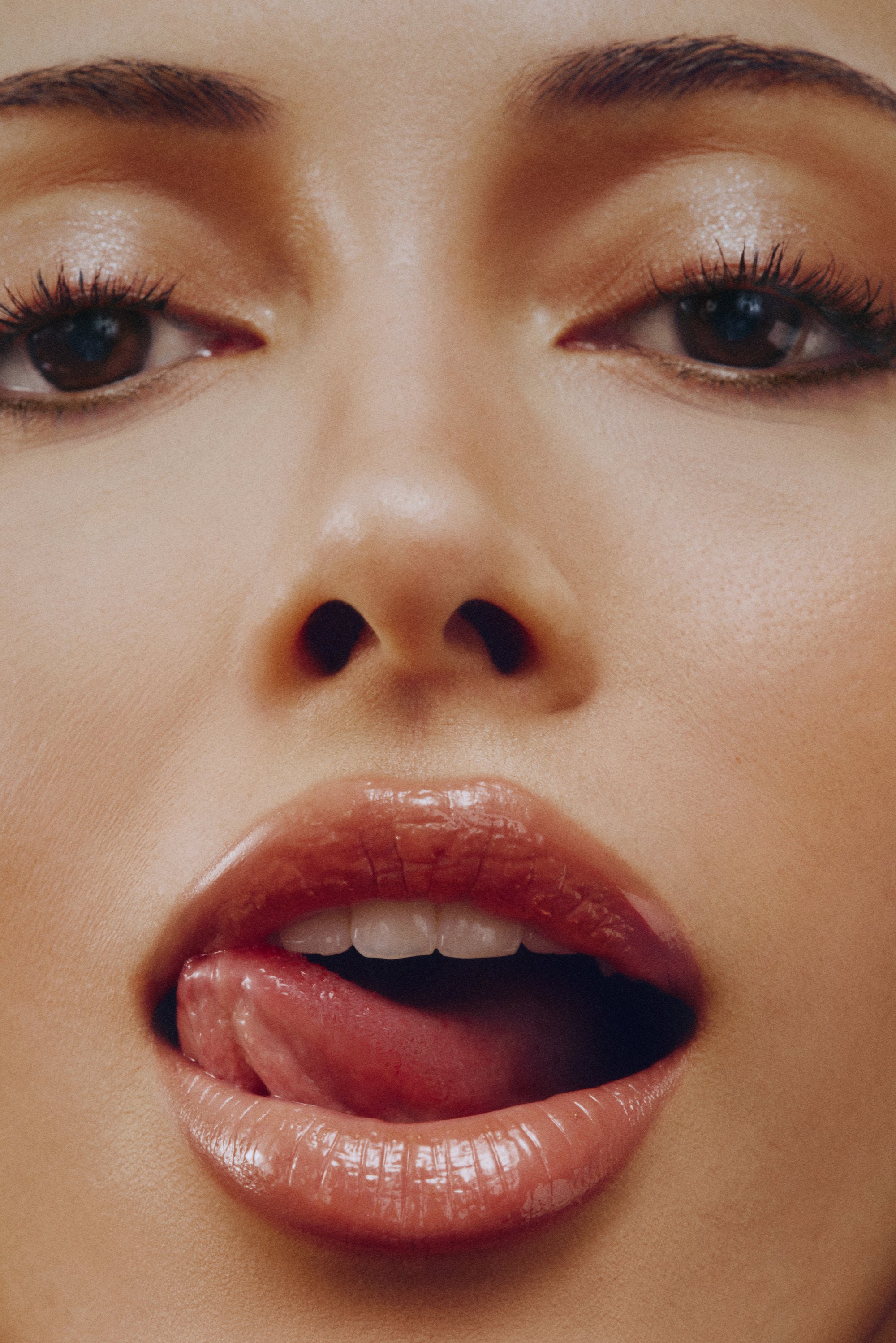 Primer plano de un rostro con labios llenos y brillantes con un tinte rojizo, ligeramente entreabiertos, con la lengua visible. Piel suave y pestañas definidas.