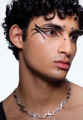 Gros plan du visage d'un modèle avec un maquillage des yeux noir complexe, trois clous en argent sur le front et un collier en chaîne argentée avec des formes abstraites.