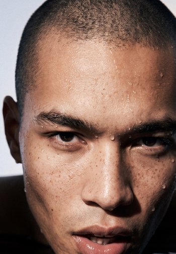 Gros plan d'un homme avec des cheveux courts et des taches de rousseur, des gouttes d'eau sur le visage, regardant directement la caméra avec une expression neutre.