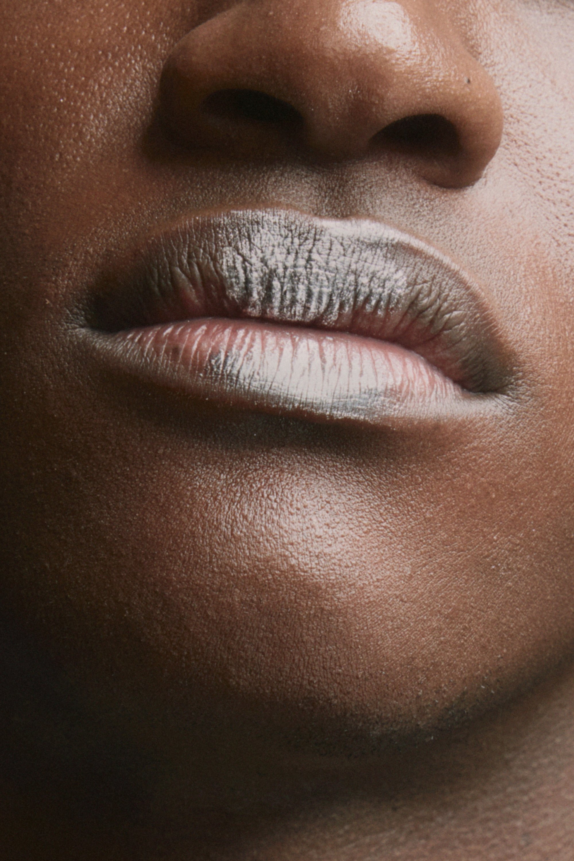 Acercamiento de la boca de una persona con labios carnosos, piel oscura lisa y sutiles destellos, contra un fondo desenfocado.