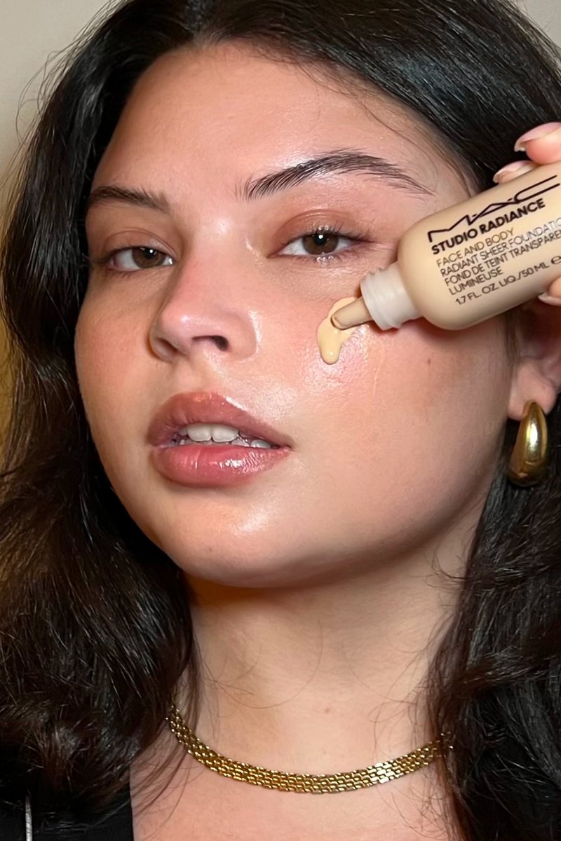 A woman with wavy hair applies a makeup serum to her cheek, holding a bottle labeled "Studio Radiance" in one hand. Soft lighting illuminates her face.