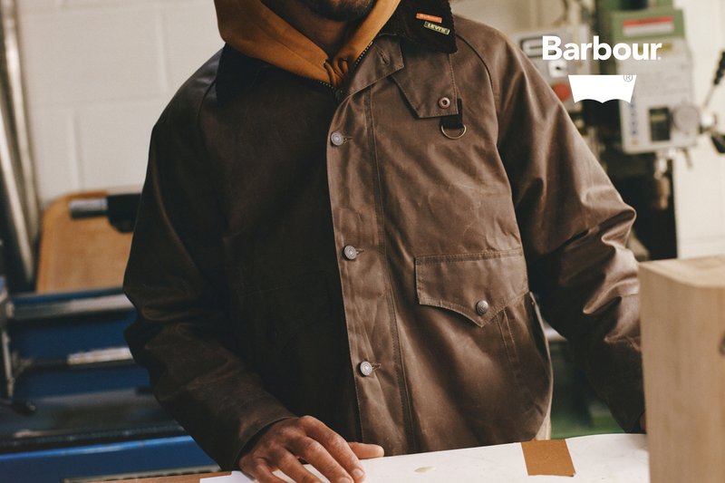 Wachsfähiger brauner Mantel mit einem Cordkragen, Druckknöpfen und Taschen, getragen über einem grauen Hoodie; Hände ruhen auf einem Arbeitstisch.