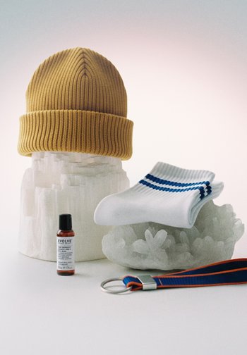 Musztardowożółta dzianinowa czapka na białym kryształowym tle, złożone białe skarpetki z niebieskimi paskami na przezroczystym kryształowym tle, mała brązowa buteleczka oraz niebiesko-pomarańczowy brelok.