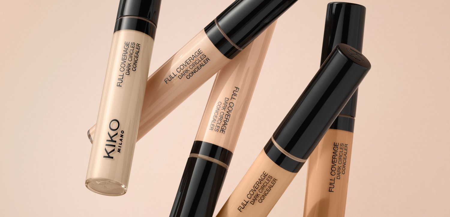 Five tubes of KIKO Milano full coverage dark circles concealer in various beige shades with black caps on a beige background.