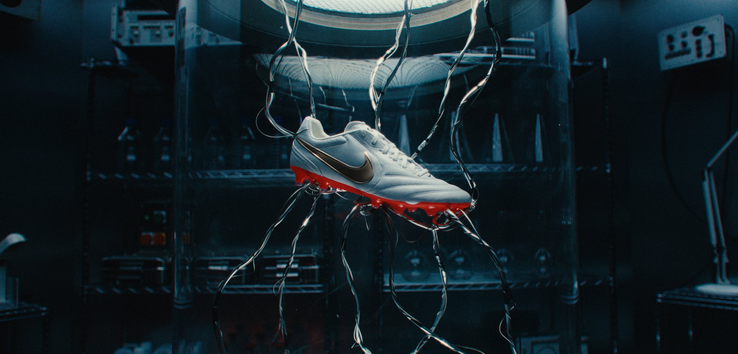 Weißer Fußballschuh mit roten Stollen und einem schwarzen Nike-Logo, der an mehreren Drähten in einer dunklen industriellen Umgebung hängt.
