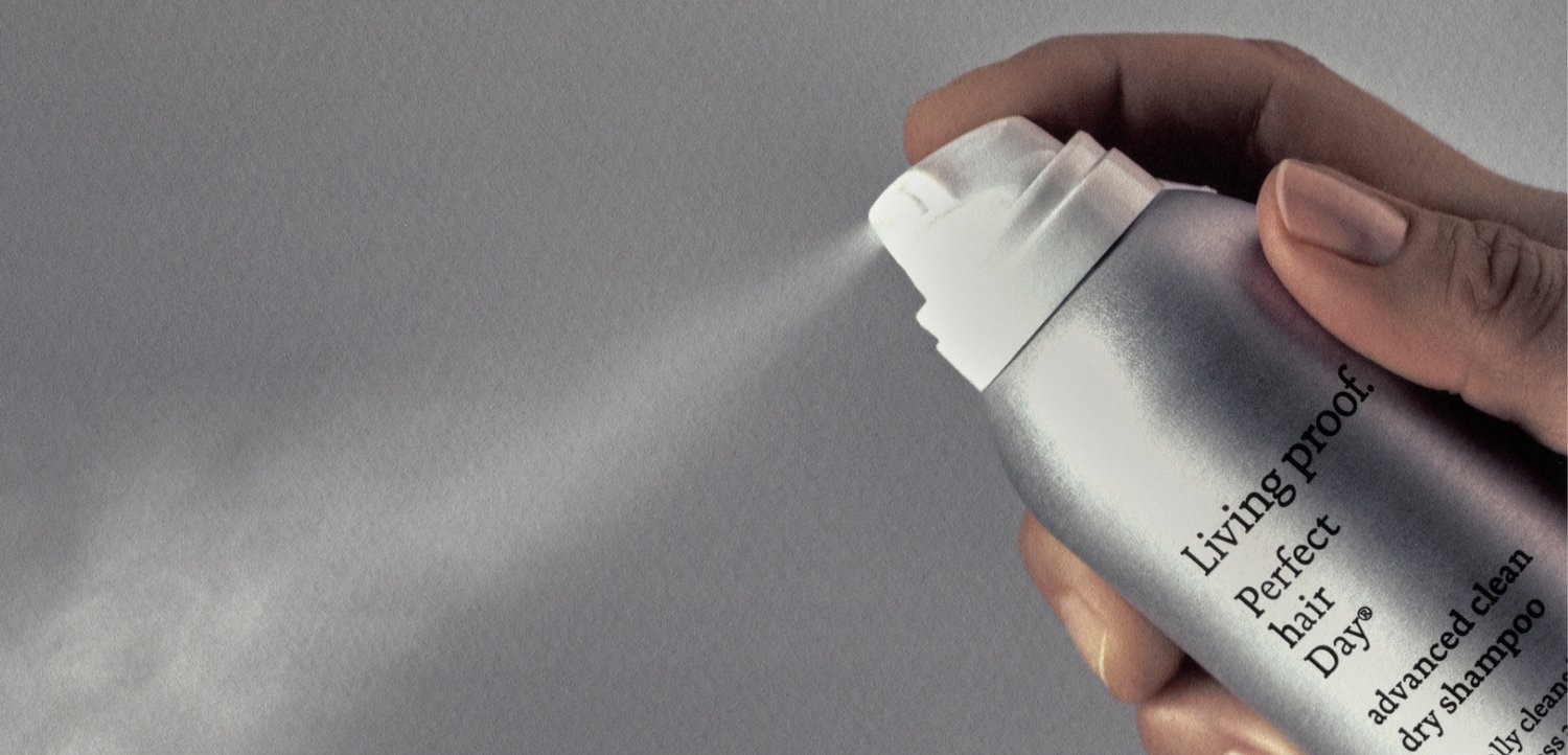 Ręka spryskuje suchy szampon Living proof Perfect Hair Day ze srebrnej puszki, uwalniając drobną mgiełkę na jednolitym szarym tle.