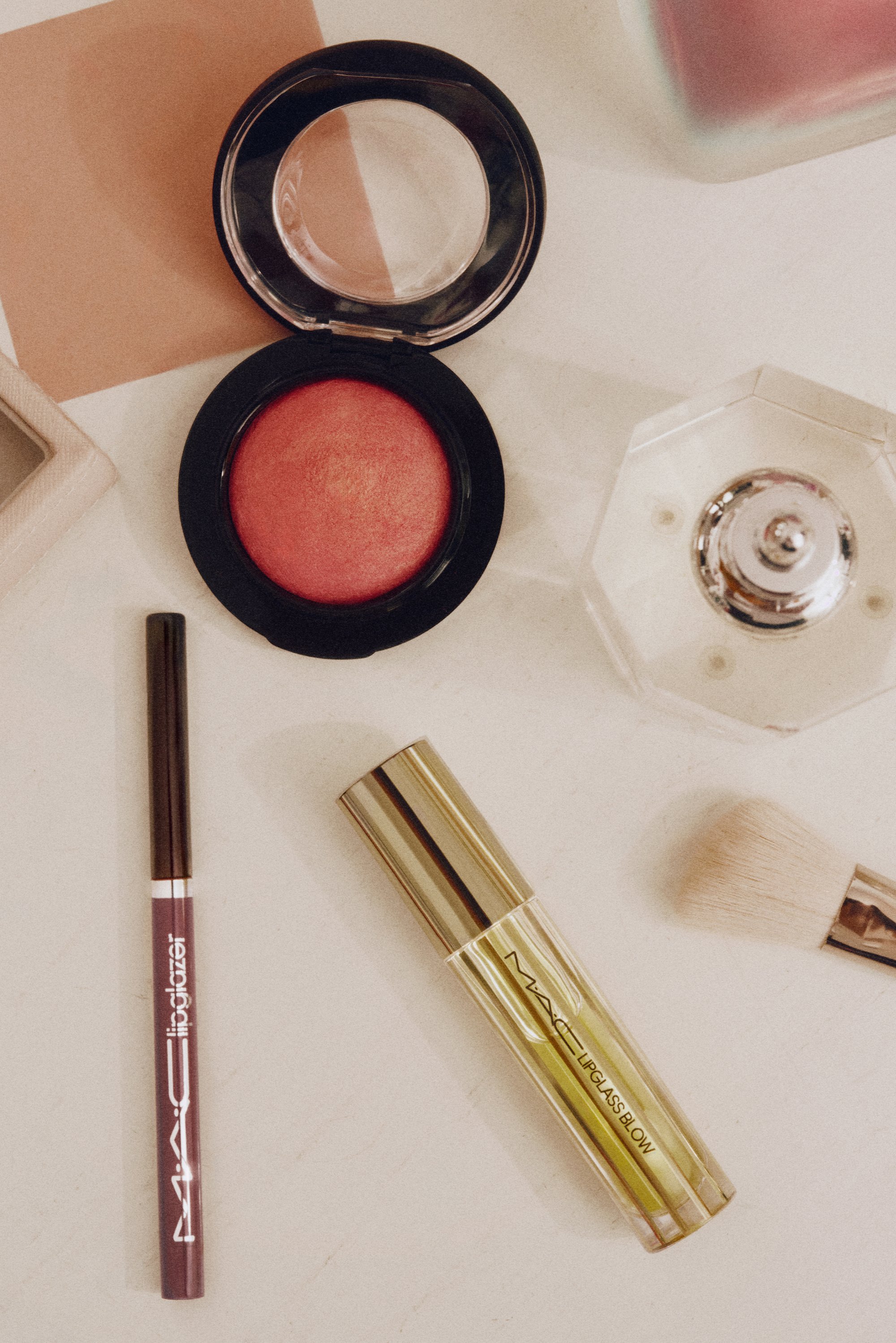 Compacto con rubor rojo, delineador de labios, brillo labial, pincel de maquillaje y botella de perfume transparente sobre superficie clara.