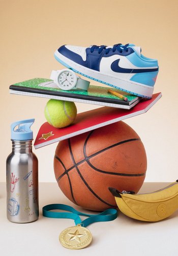 Modro-bela superga, ura, zvezki, svinčnik, teniška žogica uravnotežena na košarkarski žogi, steklenica vode, zlata medalja in rumena torbica na bež ozadju.