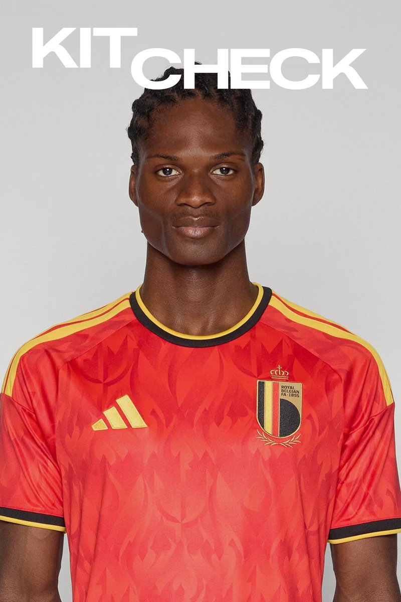 Homme aux cheveux tressés portant le maillot rouge de l'équipe nationale de football de Belgique avec des bandes jaunes et un col noir, regardant droit devant sur un fond uni.