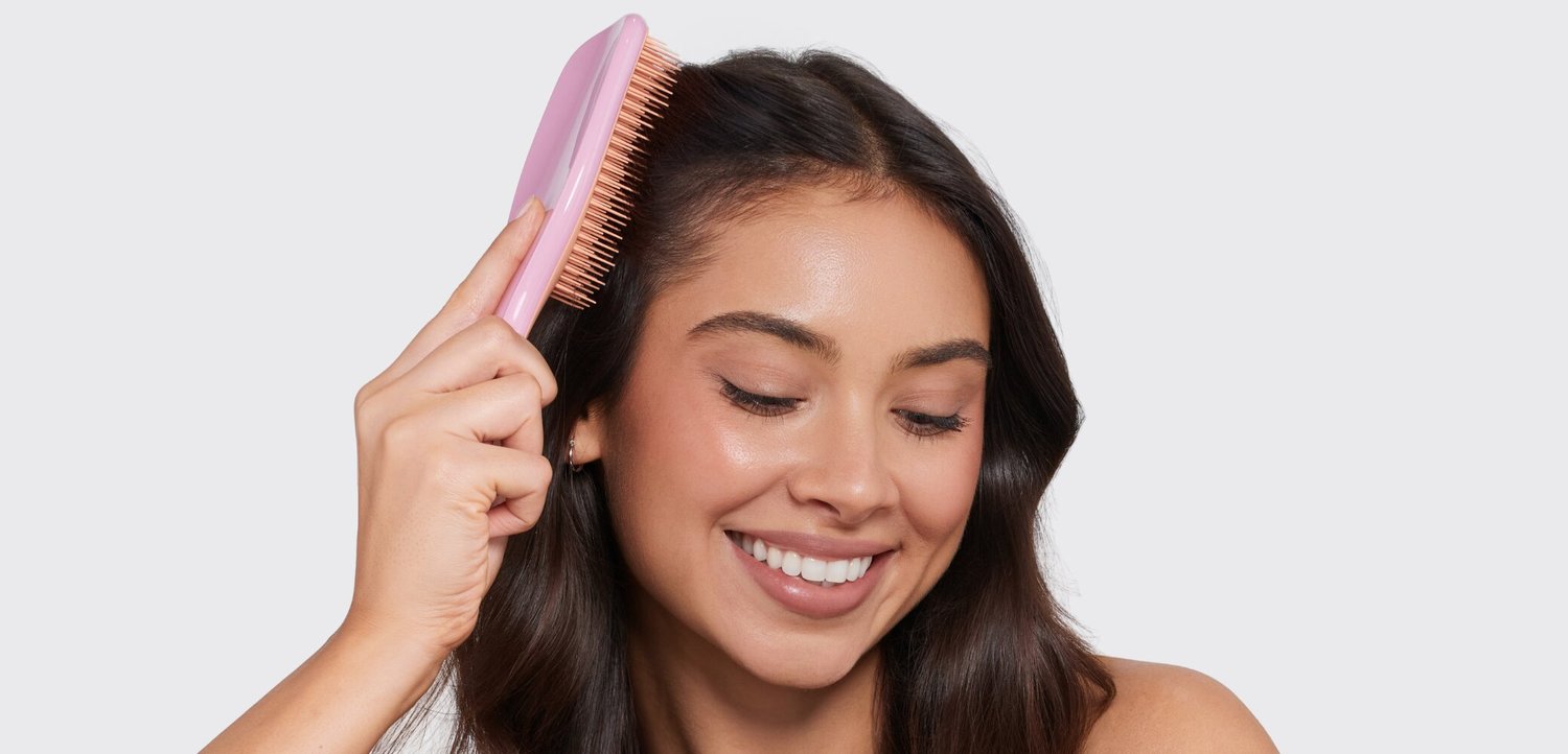 Giovane donna sorridente mentre si pettina i lunghi capelli scuri con una spazzola rosa su uno sfondo chiaro uniforme.