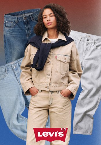 Donna con giacca di jeans beige e jeans, in piedi con le mani nelle tasche, sullo sfondo di jeans blu, bianchi e scuri e logo Levi's.