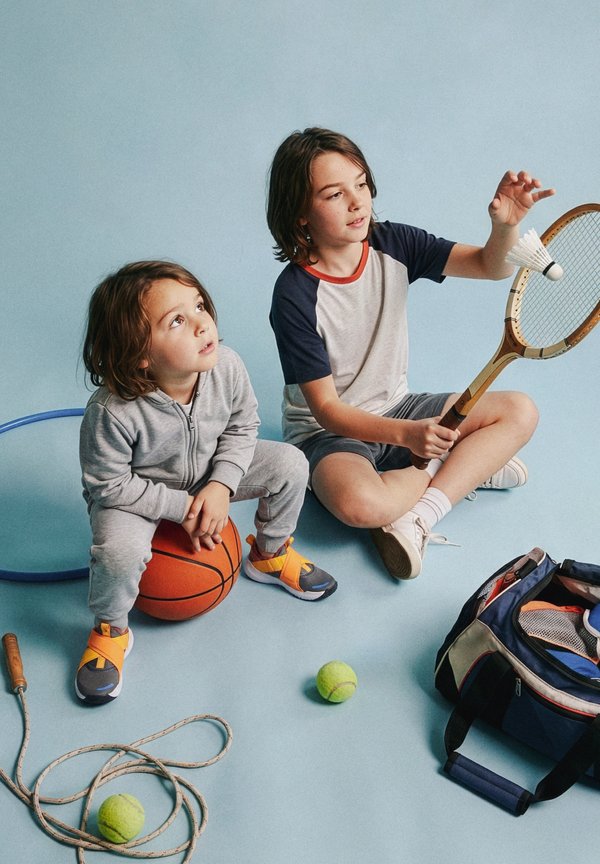 Deux enfants assis sur un sol bleu ; l'aîné tient une raquette de tennis et un volant, le plus jeune est assis sur un ballon de basket ; du matériel de sport est à proximité.