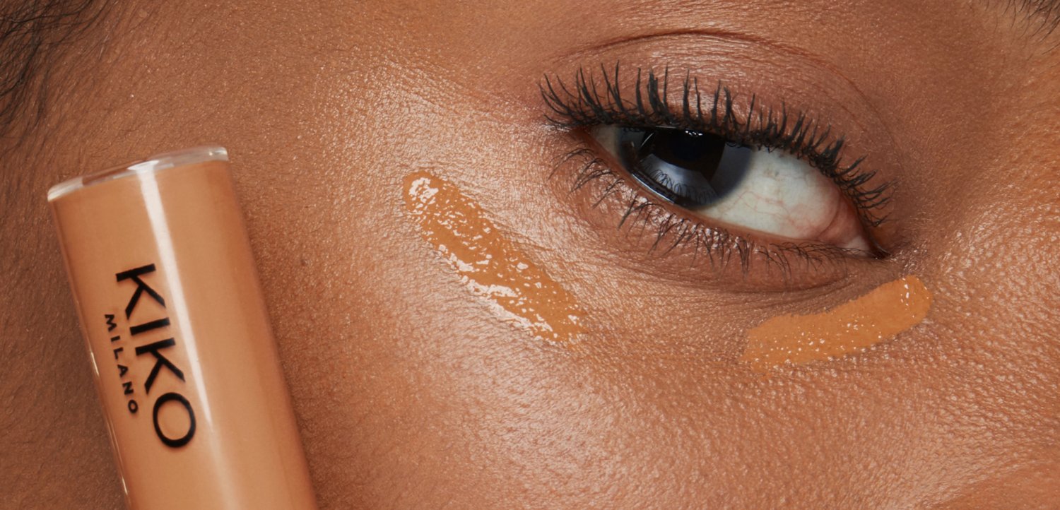Close-up of a dark-skinned face with two swatches of beige liquid concealer under the eye and a KIKO Milano concealer stick held near the cheek.