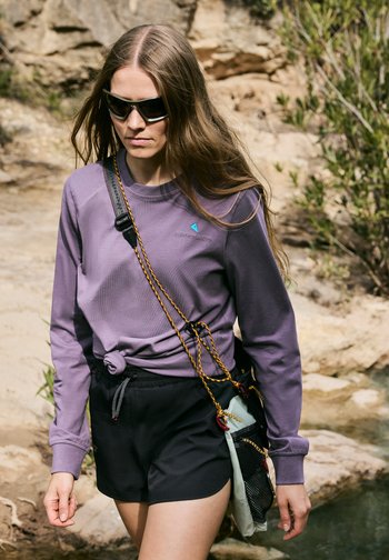 Woman with long hair wearing sunglasses, a purple sweatshirt, black shorts, and a crossbody bag, walking outdoors near rocky terrain.