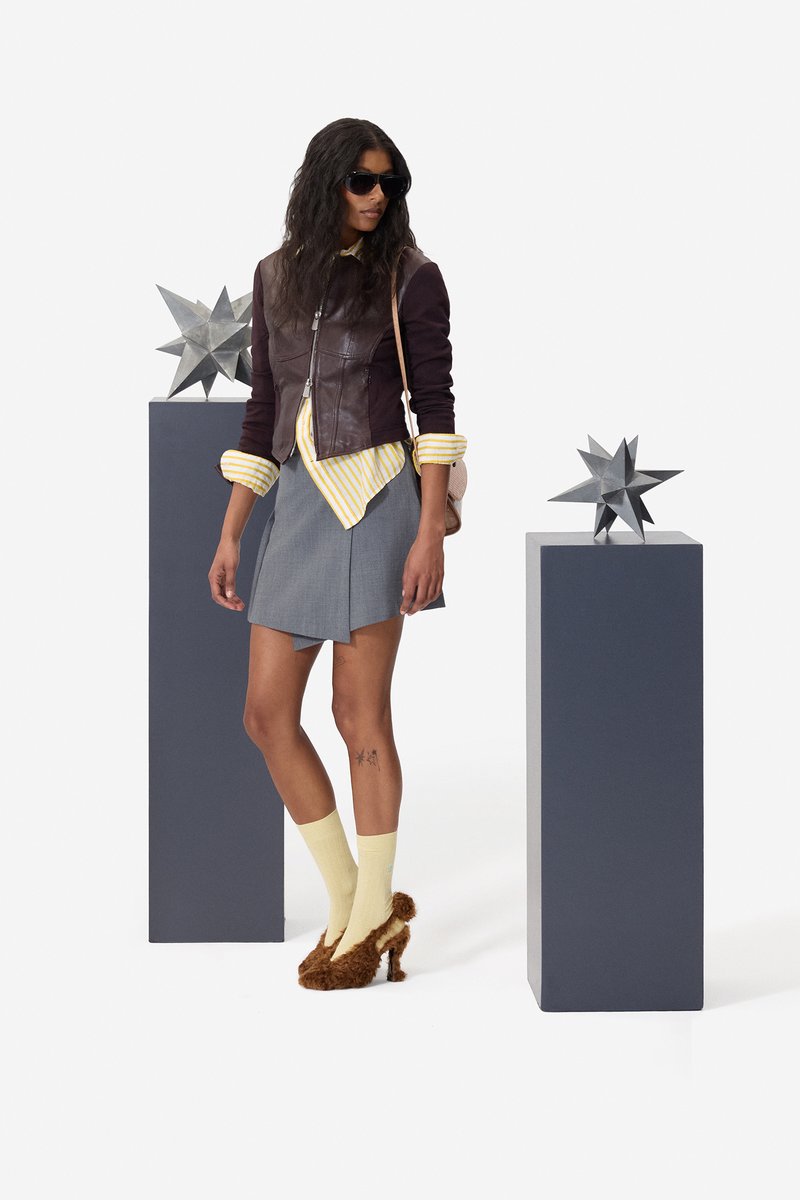 Frau mit Sonnenbrille, brauner Jacke, gelb gestreiftem Hemd, grauem Rock, gelben Socken und braunen pelzigen Absätzen steht zwischen zwei Sockeln mit Stern-Skulpturen.