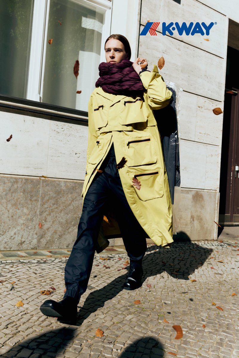 Gele waterdichte trenchcoat met meerdere zakken, gecombineerd met zwarte broek en glanzende enkellaarsjes, lopend op een kinderkopjesstraat. Paarse sjaal.