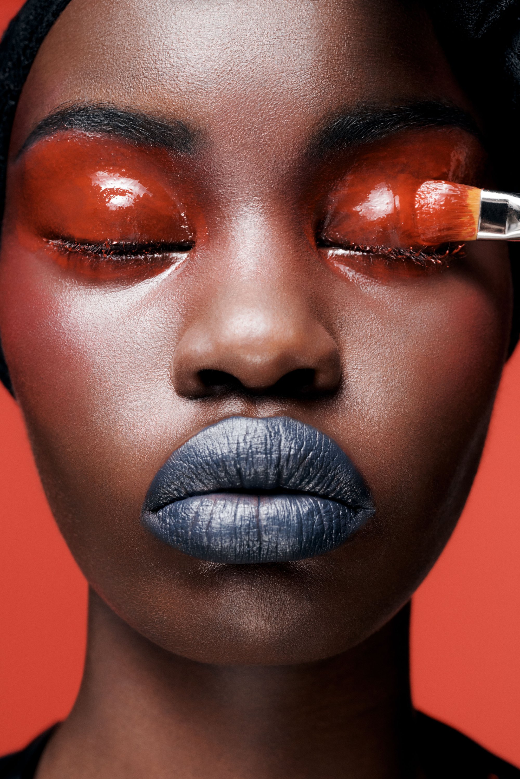 Model with bold blue lips and glossy red eyelids, eyes closed, against a vibrant orange background. A brush applies product near her eyes.