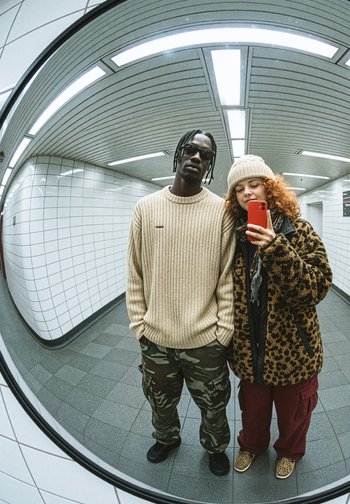 To personer i en speilrefleksjon; den ene har på seg en beige ribbestrikket genser og kamuflasjebukser, den andre har en leopardmønstret kåpe og røde bukser.