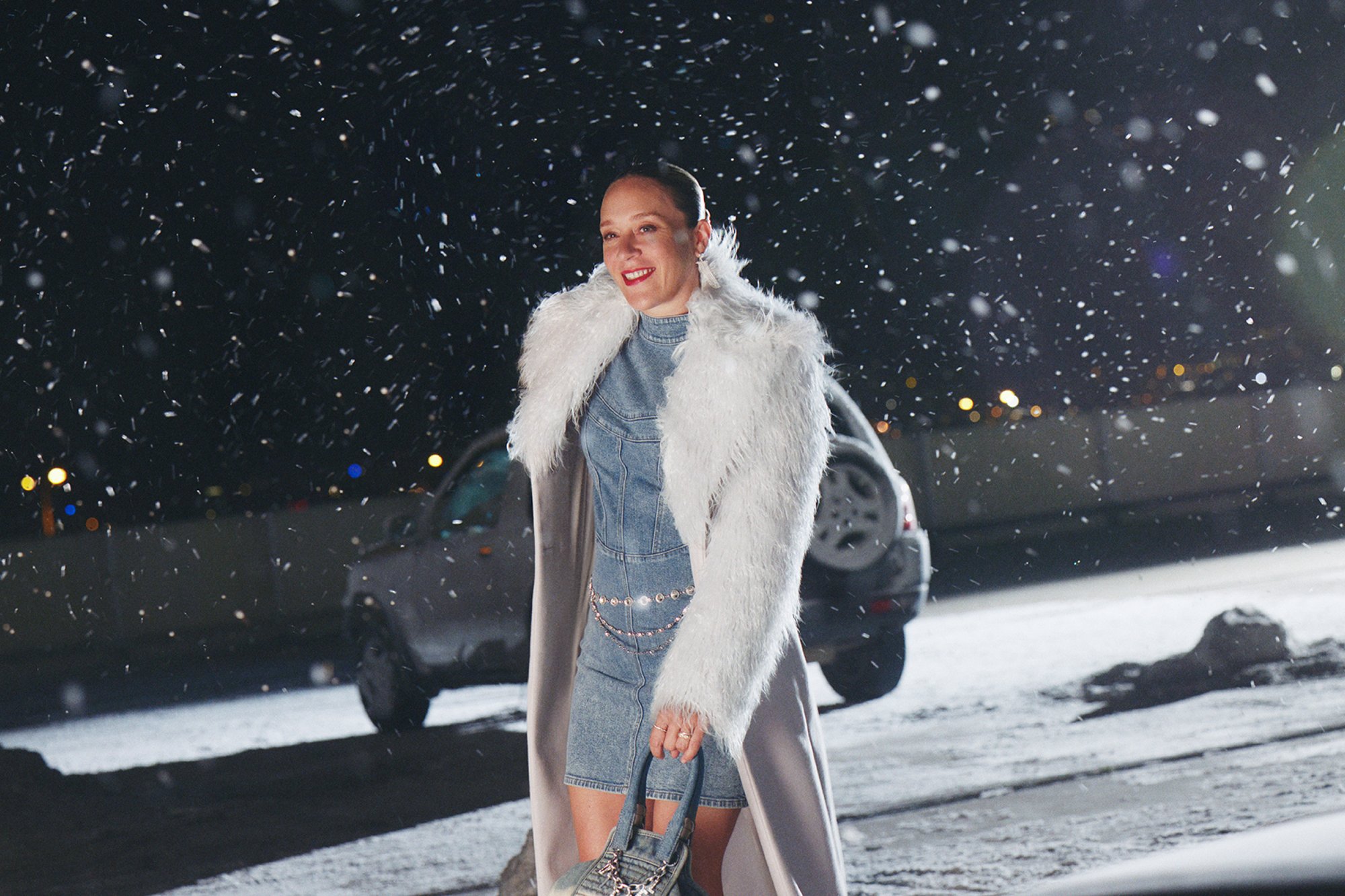 Una donna con un cappotto di pelliccia bianca e un vestito di denim sorride nella neve che cade, tenendo una borsa, con un'auto e luci della città sfocate sullo sfondo.
