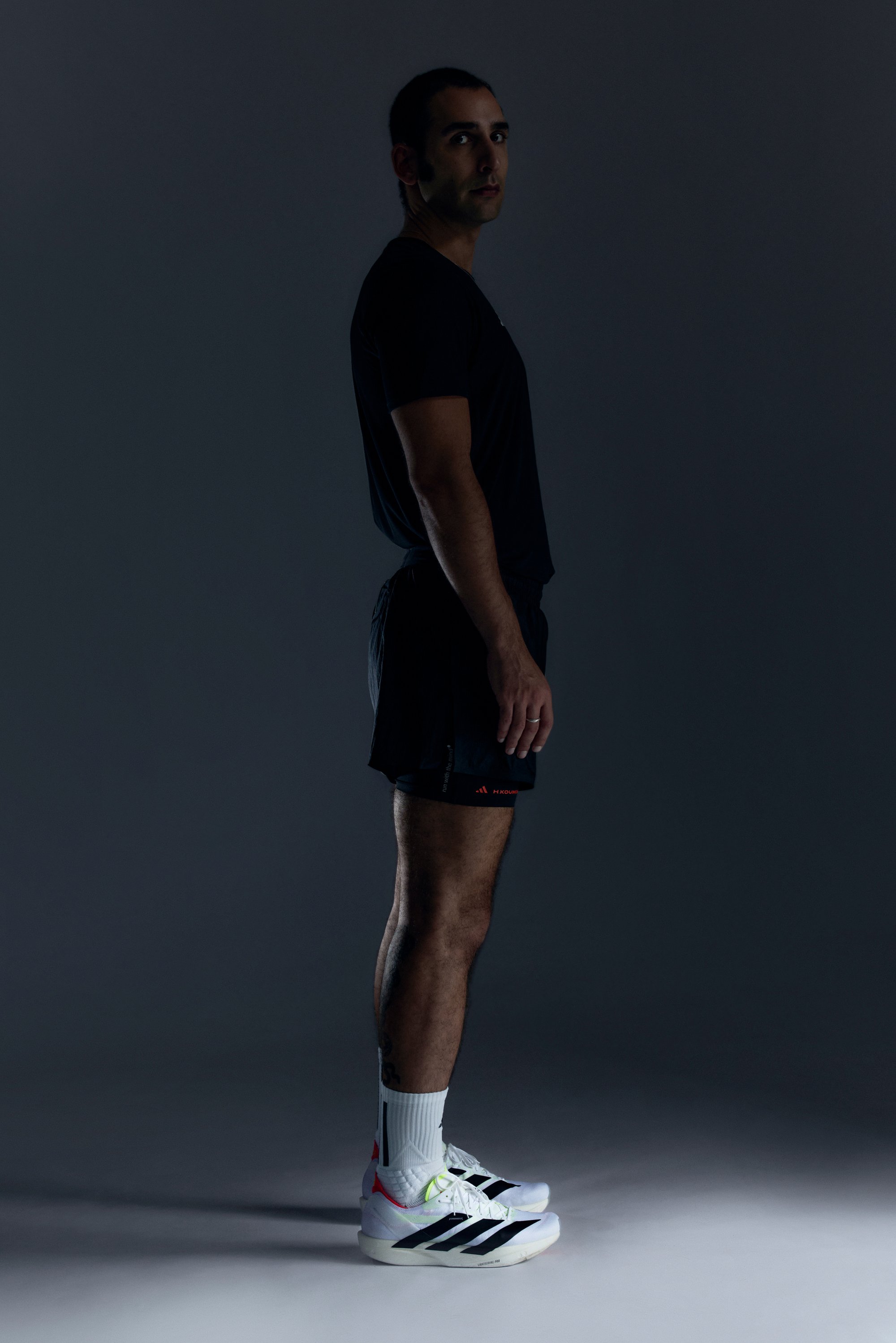 Mann in schwarzer Sportkleidung, der seitlich steht, mit weißen und schwarzen Laufschuhen und weißen Socken, vor einem dunkelgrauen Hintergrund.
