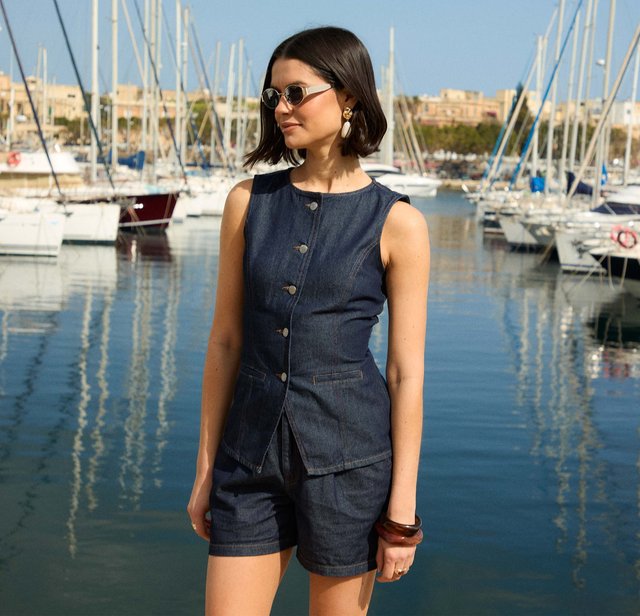 Frau trägt ärmelloses dunkles Denim-Top und Shorts und steht an einem Hafen mit Segelbooten und ruhigem Wasser an einem sonnigen Tag.