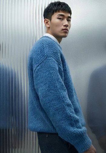 Blå strikkegenser med teksturert mønster, med lange ermer og en avslappet passform. Brukes over en hvit skjorte med krage og mørke bukser.