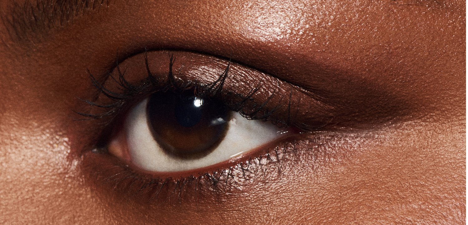 Gros plan d'un œil marron avec des cils foncés, une peau lisse et un léger brillant sur la paupière, mettant en valeur une teinte riche et texturée.