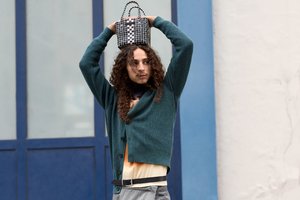 Eine Person mit lockigem Haar hält eine gemusterte Tasche über den Kopf und trägt einen grünen Cardigan über einem hellen Hemd, vor blauen und weißen Wänden.