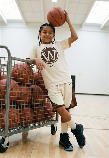 Junger Basketballspieler in Sportkleidung lehnt an einem Ballwagen und hält einen Basketball in einer Halle mit Holzfußboden hoch.