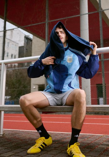 Jonge man in sportjas en korte broek zittend op een bankje bij een atletiekbaan, gezicht droogwrijvend met een donkere handdoek, gele sportschoenen zichtbaar.