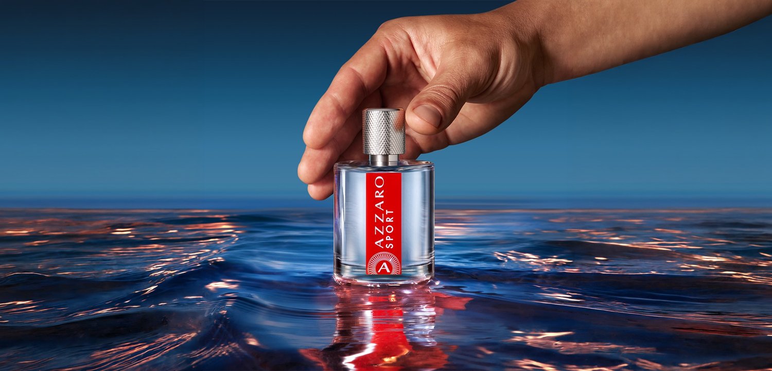 Hand holding a bottle of Azzaro Sport cologne partially submerged in calm water against a blue sky background.