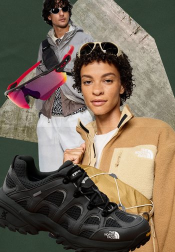Schwarze Outdoor-Schuhe mit strukturiertem Mesh und glatten Akzenten. Beiger Fleece-Jacke mit Logotasche und bunten Sonnenbrillen im Hintergrund.