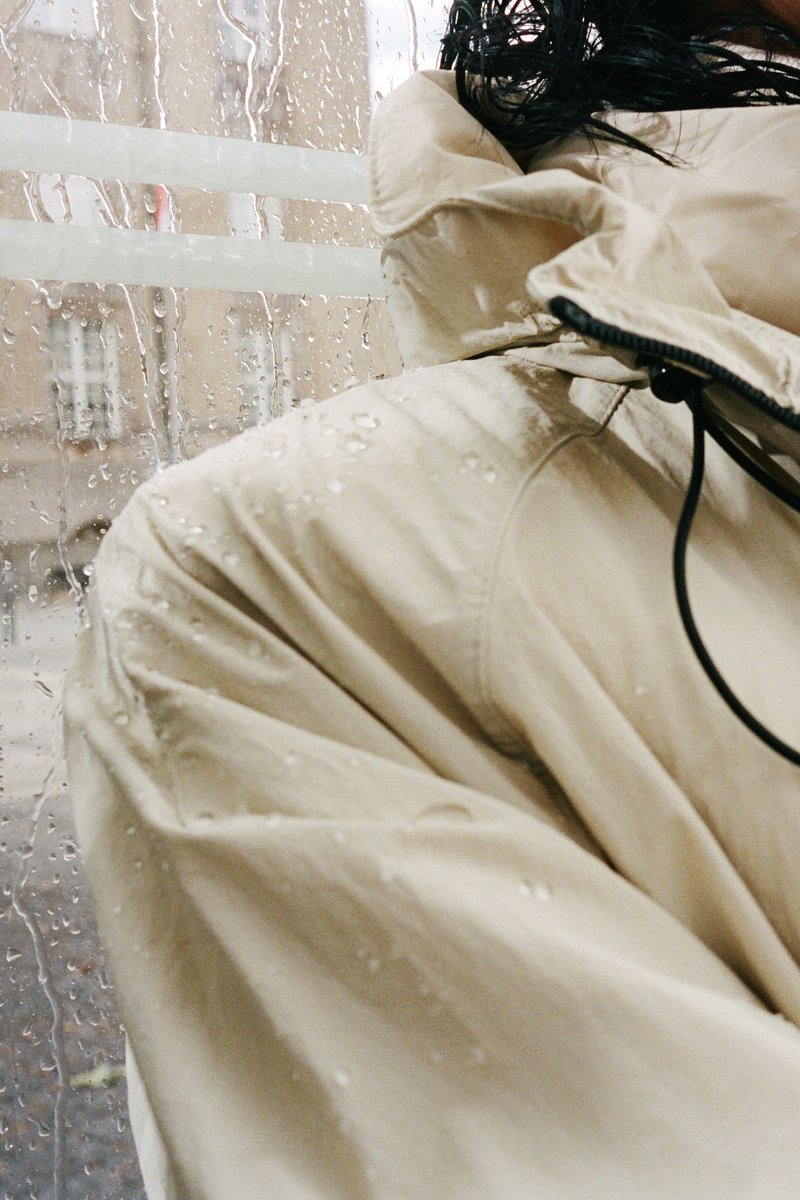 Beaded raindrops cling to a beige jacket, hinting at rain outside a window, with soft light filtering through the wet glass.