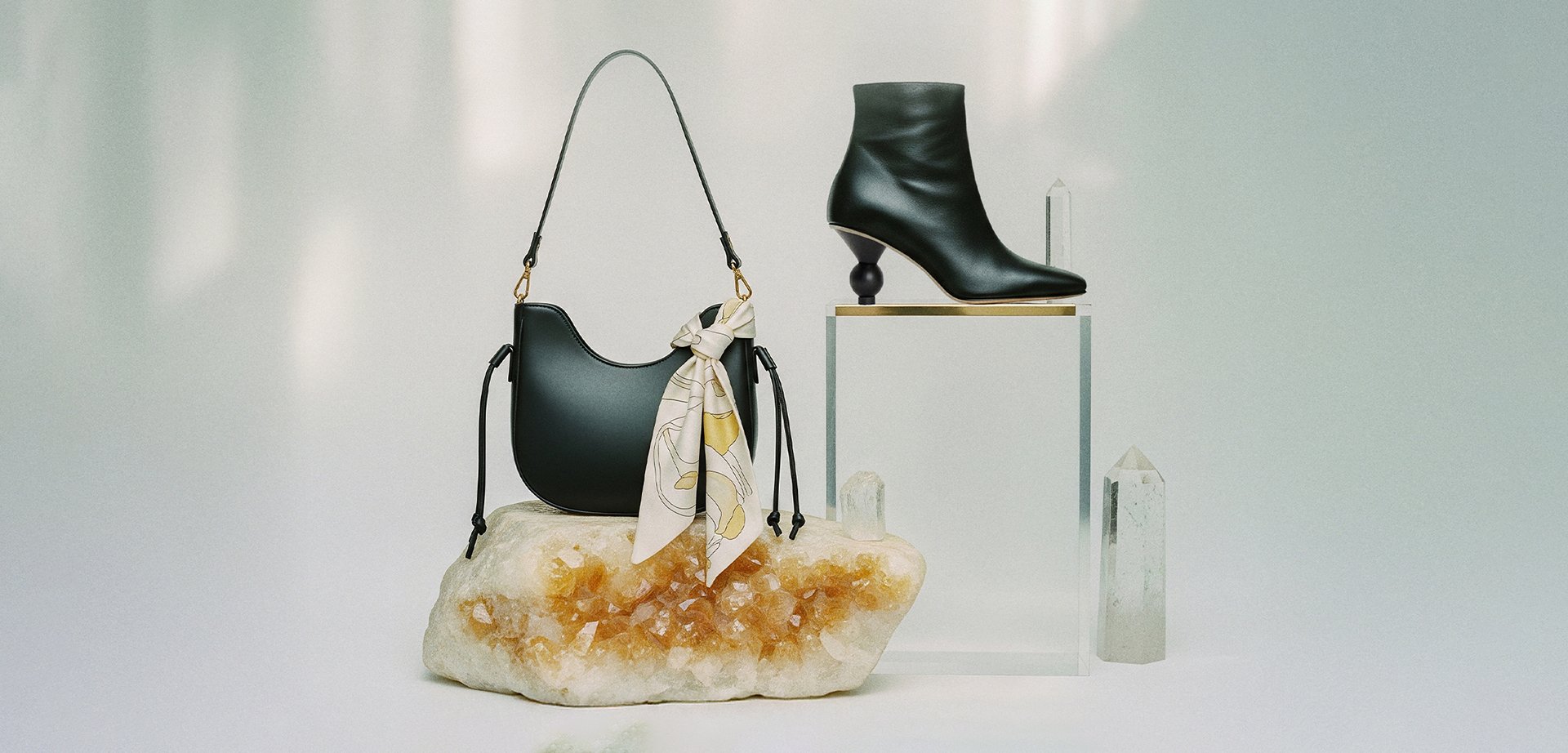 Black leather handbag adorned with a patterned scarf, next to a black ankle boot featuring a sculptural heel, placed on a textured stone base.