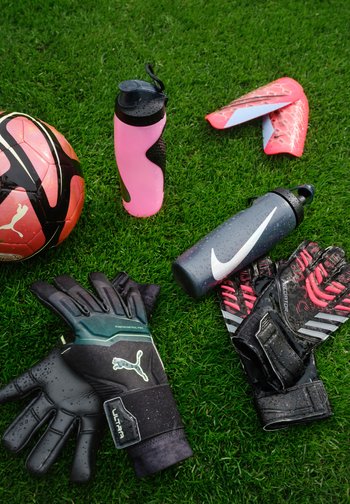 Un ballon de football Puma, deux bouteilles d'eau (rose, noir) et deux paires de gants de gardien de but noirs, ornés d'accents verts et rouges, sur de l'herbe.