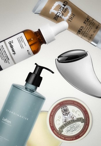 Herrenpflegeprodukte einschließlich einer Serumflasche, feuchtigkeitsspendender Lotion mit Spender, Dose mit Styling-Lehm, Tube Haar-Gel und einem silbernen Gesichtsmassager.