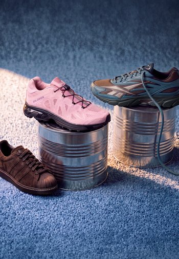 Three different sneakers displayed on and near two metal cans on blue textured ground, with one pink, one blue, and one brown shoe.