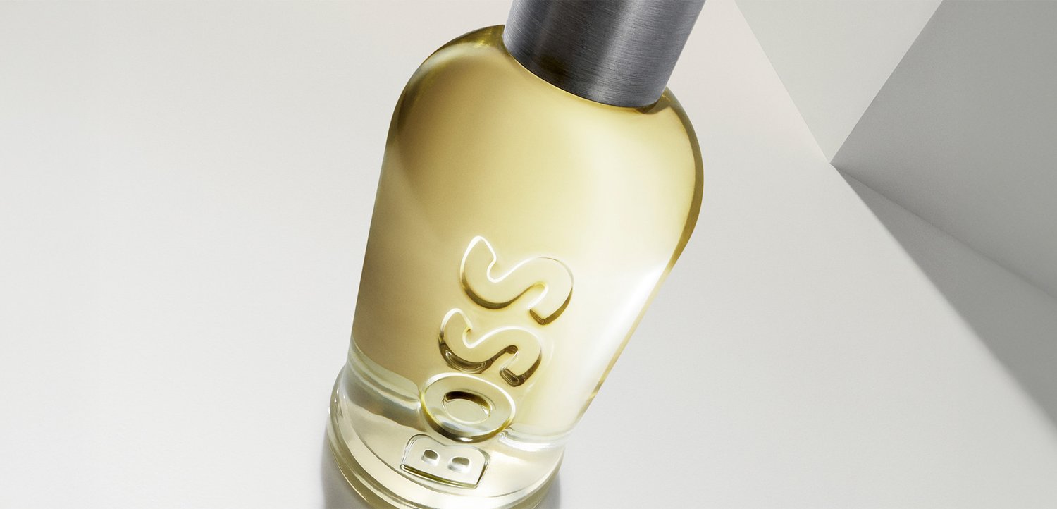 Bottiglia di profumo in vetro trasparente con liquido giallo chiaro, tappo argentato e nome del marchio in rilievo "BOSS" su una superficie bianca.