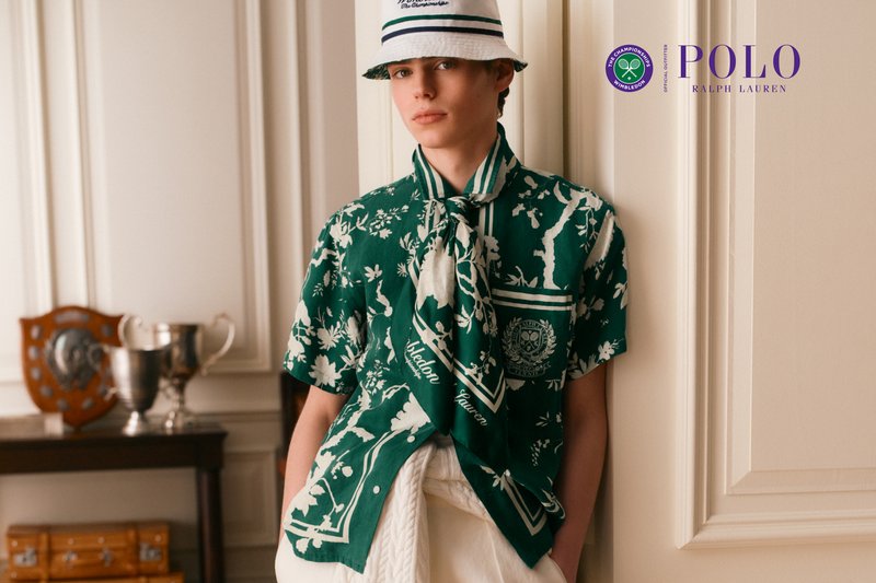 Junge Person trägt ein grünes, geblümtes Polo Ralph Lauren Hemd und einen weißen Bucket Hat, steht drinnen in der Nähe von Trophäen und Auszeichnungen.