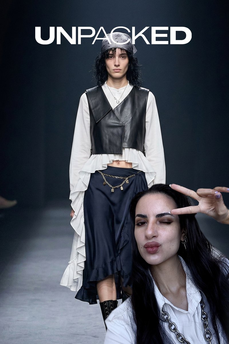 Frau in geschichtetem schwarz-weißem Outfit mit Kettengürtel geht hinter einer anderen Frau entlang des Laufstegs, die ein Friedenszeichen macht und die Lippen spitzt.